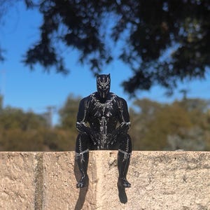 May include: A black figurine of a superhero character with a pointed mask and silver-coloured detailing on the chest, arms, and legs. The figure is depicted in a seated position with hands resting on the knees, showcasing a textured, moulded surface.
