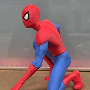 May include: A red, blue, and black Spider-Man figurine in a crouching pose. The figure is wearing a red and black mask with white eye lenses, a red torso, blue arms and legs, and red gloves and boots. The figure is kneeling on one knee with one arm extended.
