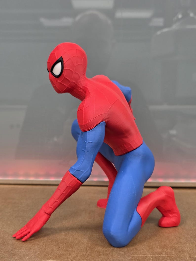 May include: A red, blue, and black Spider-Man figurine in a crouching pose. The figure is wearing a red and black mask with white eye lenses, a red torso, blue arms and legs, and red gloves and boots. The figure is kneeling on one knee with one arm extended.