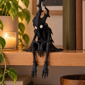Könnte beinhalten: Eine mattschwarze, bewegliche Figur mit einem schlanken, gezackten, baumartigen Kopf und langen, klauenartigen Gliedmaßen. Die Figur hat zwei große, kreisförmige, leuchtend weiße Augen und sitzt in einer Position mit herabhängenden Beinen.