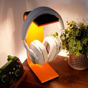 May include: White over-ear headphones on an orange and black headphone stand. A green and black game controller is on a wooden surface, with a potted plant in the background. The stand has a curved design.