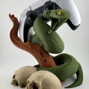 Może przedstawiać: Zielona rzeźba węża z kontrolerem PlayStation 5 spoczywającym na brązowej gałęzi. Dwie czaszki znajdują się u podstawy. Wąż ma żółte oczy i otwartą paszczę. Kontroler jest biało-czarny.