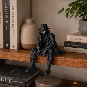 May include: A black figurine of a character wearing a fedora, a long trench coat, and boots, depicted in a seated position with legs dangling. The figure features white eye lenses and detailed clothing textures, crafted in a monochromatic style.