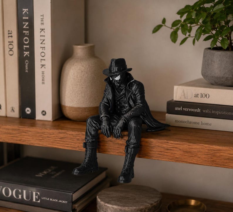 May include: A black figurine of a character wearing a fedora, a long trench coat, and boots, depicted in a seated position with legs dangling. The figure features white eye lenses and detailed clothing textures, crafted in a monochromatic style.