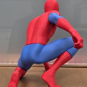 May include: A red, blue, and black Spider-Man figurine in a crouching pose. The figure is wearing a red suit with blue accents on the arms and legs. The figurine is a detailed representation of the superhero.