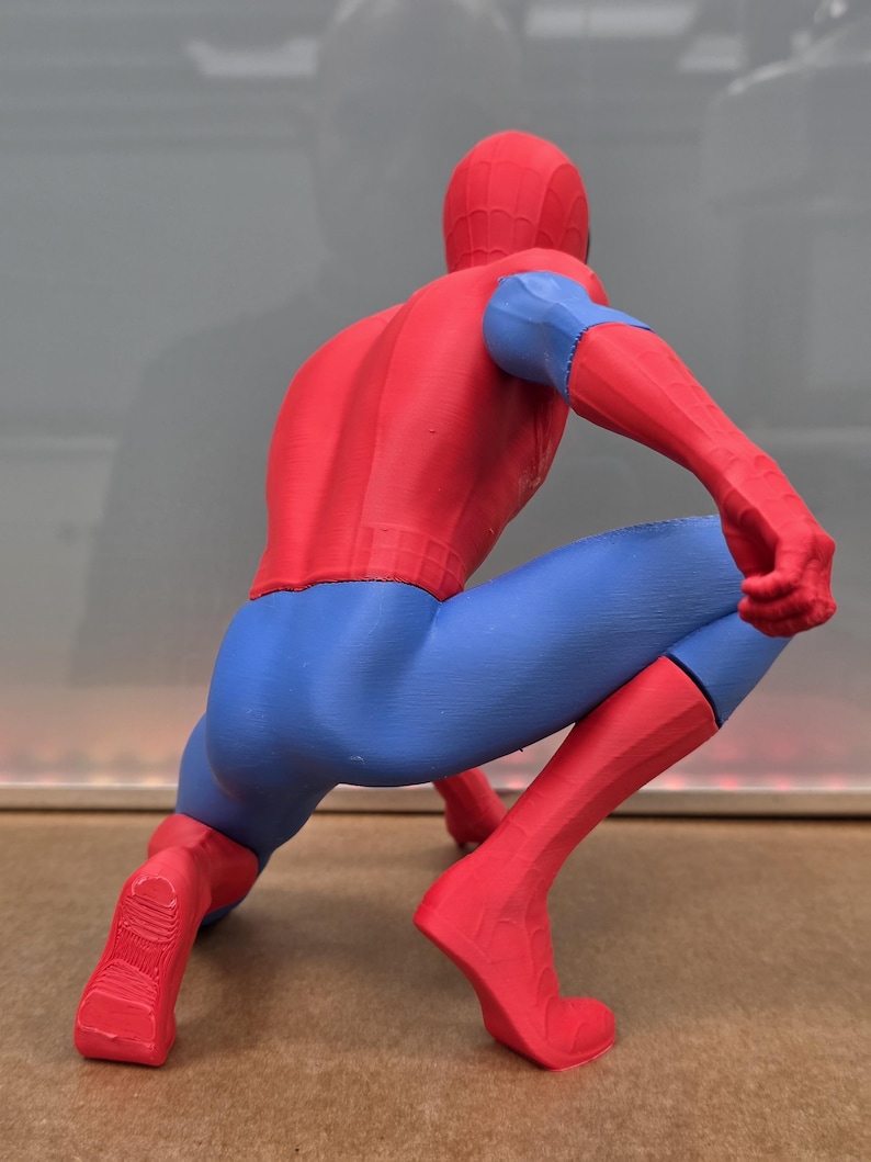 May include: A red, blue, and black Spider-Man figurine in a crouching pose. The figure is wearing a red suit with blue accents on the arms and legs. The figurine is a detailed representation of the superhero.