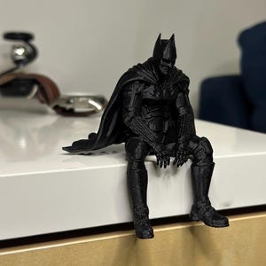 以下が含まれることがあります： マットブラックの3Dプリント製バットマンのフィギュア。縁から足をぶら下げて座っているポーズで、詳細なマント、アーマースーツの質感、象徴的な尖ったカウルが特徴です。全体が単一の暗い色で表現されています。