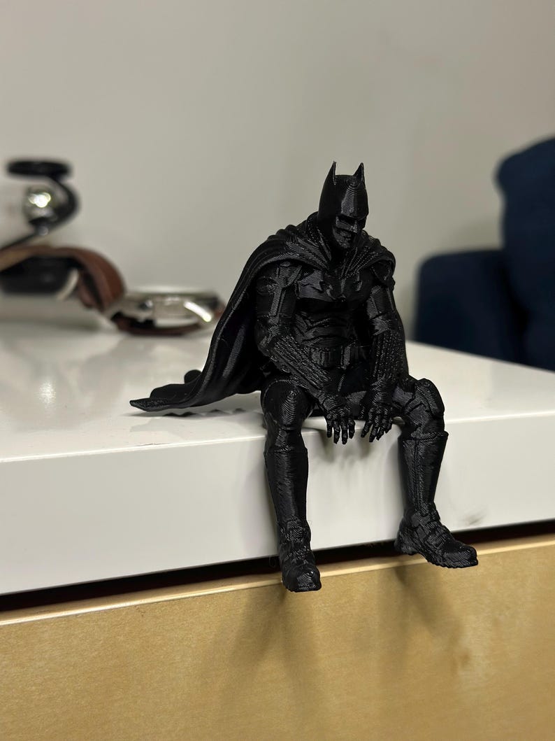 以下が含まれることがあります： マットブラックの3Dプリント製バットマンのフィギュア。縁から足をぶら下げて座っているポーズで、詳細なマント、アーマースーツの質感、象徴的な尖ったカウルが特徴です。全体が単一の暗い色で表現されています。