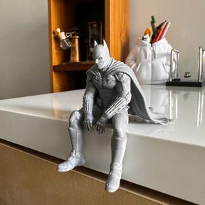 以下が含まれることがあります： 表面の端に座っている、マントを羽織ったスーパーヒーローのグレーの3Dプリントフィギュア。フィギュアには詳細なマスク、腕と脚のテクスチャ加工されたアーマー、そして背後に伸びる流れるようなマントがあり、積層痕がはっきりと見えます。