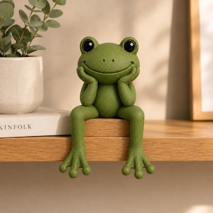 May include: A green frog figurine with a textured surface, large black eyes, and a wide smile. The frog is depicted in a seated position with its hands resting under its chin, designed to sit on the edge of a flat surface like a shelf or table.