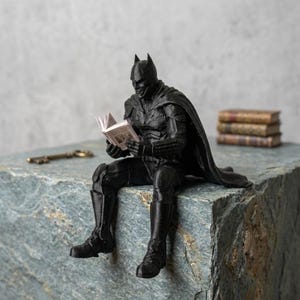 以下が含まれることがあります： 小さな開いた本を手に持ち読んでいる、マントを羽織った黒いスーパーヒーローのフィギュア。石の縁に座っており、尖ったマスクとマントが付いた質感のあるスーツを着用している。石の表面には小さな金属製の鍵が置かれている。