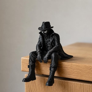 May include: A black figurine of a character wearing a fedora, a long trench coat, gloves, and tall lace-up boots. The figure is posed in a seated position with legs dangling, featuring detailed textures on the clothing and a mask covering the face.