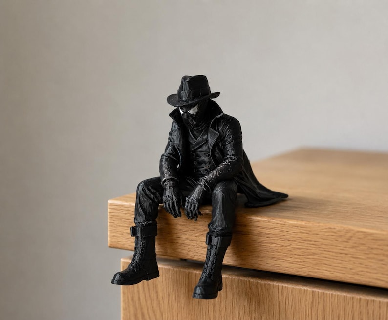 May include: A black figurine of a character wearing a fedora, a long trench coat, gloves, and tall lace-up boots. The figure is posed in a seated position with legs dangling, featuring detailed textures on the clothing and a mask covering the face.