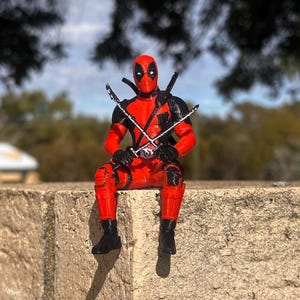Op de afbeelding: Een rood met zwart Deadpool-actiefiguur in zittende houding, voorzien van een masker met witte oogvlakken, een riem en twee gekruiste zwaarden voor de borst. Het figuur heeft zwarte laarzen, zwarte handschoenen en zwarte schouderaccenten.