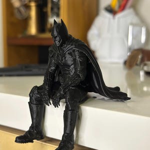 以下が含まれることがあります： 座った姿勢のケープをまとったスーパーヒーローの黒い3Dプリントフィギュア。フィギュアは詳細なマスク、胸と腕のテクスチャ加工されたアーマー、そして背後に伸びる流れるようなケープが特徴です。表面には積層痕が見られます。