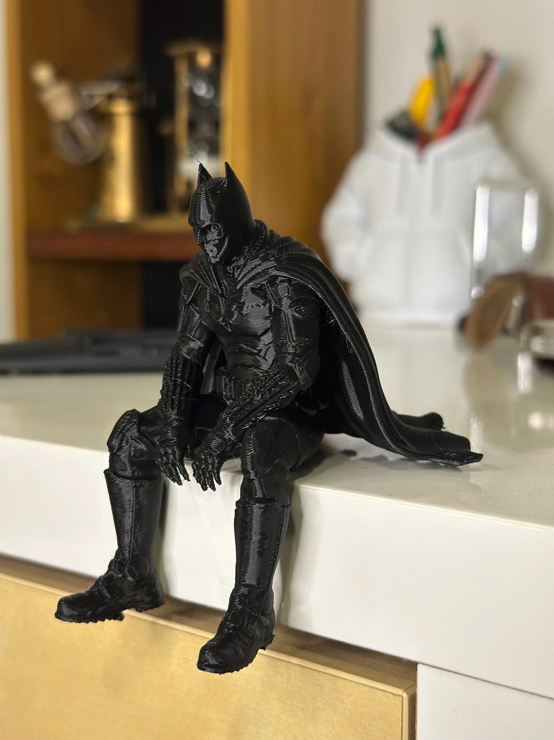以下が含まれることがあります： 座った姿勢のケープをまとったスーパーヒーローの黒い3Dプリントフィギュア。フィギュアは詳細なマスク、胸と腕のテクスチャ加工されたアーマー、そして背後に伸びる流れるようなケープが特徴です。表面には積層痕が見られます。