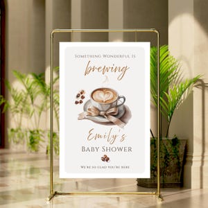May include: A gold metal frame holds a sign with the text "Something Wonderful Is Brewing" above a watercolor image of a coffee cup with a heart-shaped design. The sign reads "Emily's Baby Shower" and "We're so glad you're here."