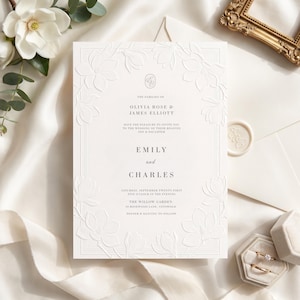 May include: A white wedding invitation card featuring an embossed floral border of magnolia flowers. The text reads: The families of Olivia Rose & James Elliott have the pleasure to invite you to the wedding of their beloved son & daughter Emily and Charles, Saturday, September twenty-first, five o'clock in the evening, The Willow Garden, 18 Rosewood Lane, Cotswold, dinner & dancing to follow.
