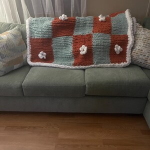 May include: A handmade, chunky knit blanket in a checkerboard pattern of rust and seafoam green. White flower appliqués are evenly spaced across the blanket. The blanket is trimmed with a thick white border and rests on a green sofa.