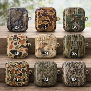 May include: Nine camouflage-patterned AirPod cases in various designs, including brown, green, and tan. Each case has a small metal loop and a unique pattern, with initials printed on each case. The cases are displayed on a wooden surface.