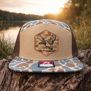 May include: A brown and tan trucker hat with a camouflage pattern on the brim and crown. The front panel features a hexagon patch with a duck graphic. The hat has a brown mesh back and a small rectangular tag with the brand name.