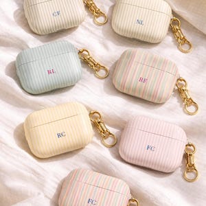 May include: Seven pastel-colored AirPod cases with vertical striped patterns and gold-colored keychains. Each case has a different initial monogram in a contrasting color. The cases are in shades of cream, blue, yellow, pink, and green.