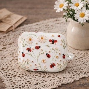May include: White case decorated with daisies and red ladybugs. The case has a clear loop for attaching to a keychain. Daisies and ladybugs are scattered across the case, with a vase of daisies in the background.