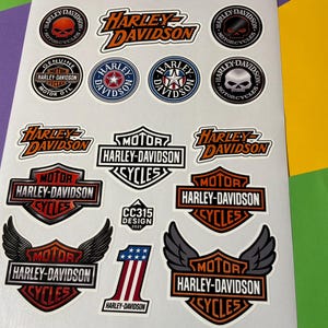 Könnte beinhalten: Ein Bogen mit Harley-Davidson-Aufklebern mit verschiedenen Logos und Designs. Die Aufkleber zeigen den Namen Harley-Davidson, Schädelgrafiken und die Nummer 1. Die Aufkleber sind in Rot, Schwarz, Weiß und Blau.