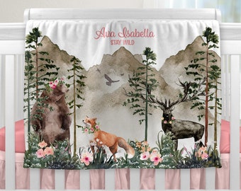 May include: A pink and white baby blanket with a woodland theme. The blanket features a bear, a fox, and a deer in a forest setting with mountains in the background. The text "Ava Isabella Stay Wild" is printed on the blanket.