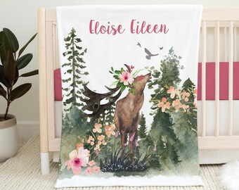 May include: A white baby blanket with a watercolor illustration of a deer in a forest setting. The deer is wearing a floral crown and there are birds flying above. The text "Eloise Eileen" is printed in pink at the top of the blanket.