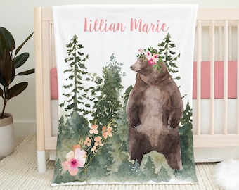 May include: A white baby blanket with a watercolor illustration of a brown bear wearing a pink flower crown. The bear is standing in a forest with green trees and pink flowers. The blanket has the name "Lillian Marie" written in pink letters.