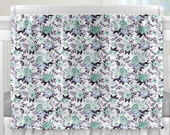 May include: A white cot with a white frilled skirt and a white and green floral baby blanket with the name "Hazel" repeated throughout the pattern.