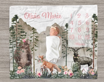 May include: A white baby blanket with a woodland theme. The blanket features a watercolor illustration of a bear, a fox, and a deer. The blanket also has a pink and white number chart for tracking a baby's growth. The text "Olivia Marie" is written in pink at the top of the blanket.