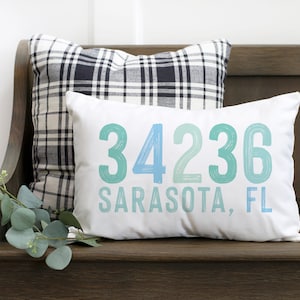 May include: White cushion with blue and green text that reads "34236 Sarasota, FL". The cushion is sitting on a wooden bench with a black and white checked cushion behind it.