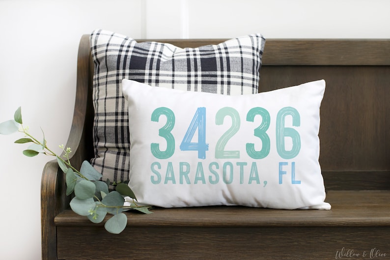 May include: White cushion with blue and green text that reads "34236 Sarasota, FL". The cushion is sitting on a wooden bench with a black and white checked cushion behind it.