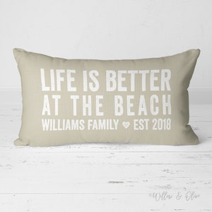 May include: Beige throw pillow with the text "Life is better at the beach Williams Family Est 2018" in white.