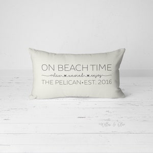Puede incluir: Almohada rectangular blanca con texto negro que dice "On Beach Time relax unwind enjoy The Pelican Est. 2016".