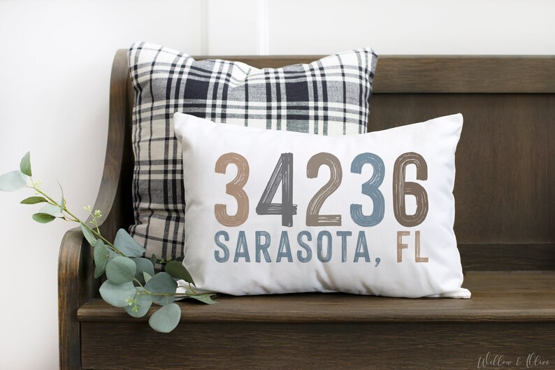 May include: White throw pillow with the text "34236 Sarasota, FL" in brown and blue lettering. The pillow is sitting on a wooden bench with a black and white plaid pillow behind it.