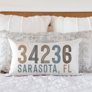 May include: A white pillow with the text "34236 Sarasota, FL" in brown, black, and blue lettering. The pillow is on a white bed with a white headboard.