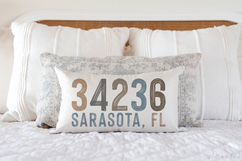 May include: A white pillow with the text "34236 Sarasota, FL" in brown, black, and blue lettering. The pillow is on a white bed with a white headboard.