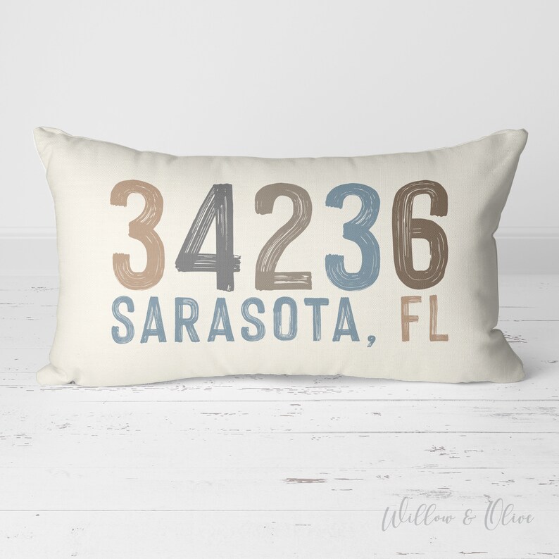 May include: A beige pillow with the text "34236 Sarasota, FL" in brown, gray, and blue brushstrokes.
