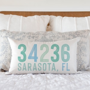 May include: A white pillow with blue and green text that reads "34236 Sarasota, FL". The pillow is on a white bed with white pillows behind it.