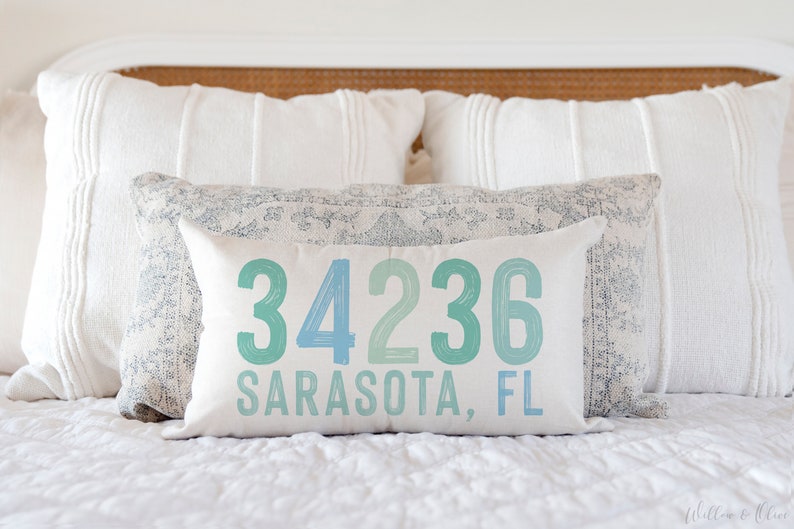 May include: A white pillow with blue and green text that reads "34236 Sarasota, FL". The pillow is on a white bed with white pillows behind it.