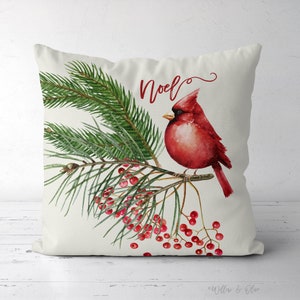 May include: A white throw pillow with a red cardinal perched on a green pine branch with red berries. The text "Noel" is written in red cursive on the pillow.