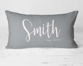 May include: A rectangular grey cushion featuring the name "Smith" in white script. Below the name is "est. 2019". The cushion is placed on a white wooden surface.