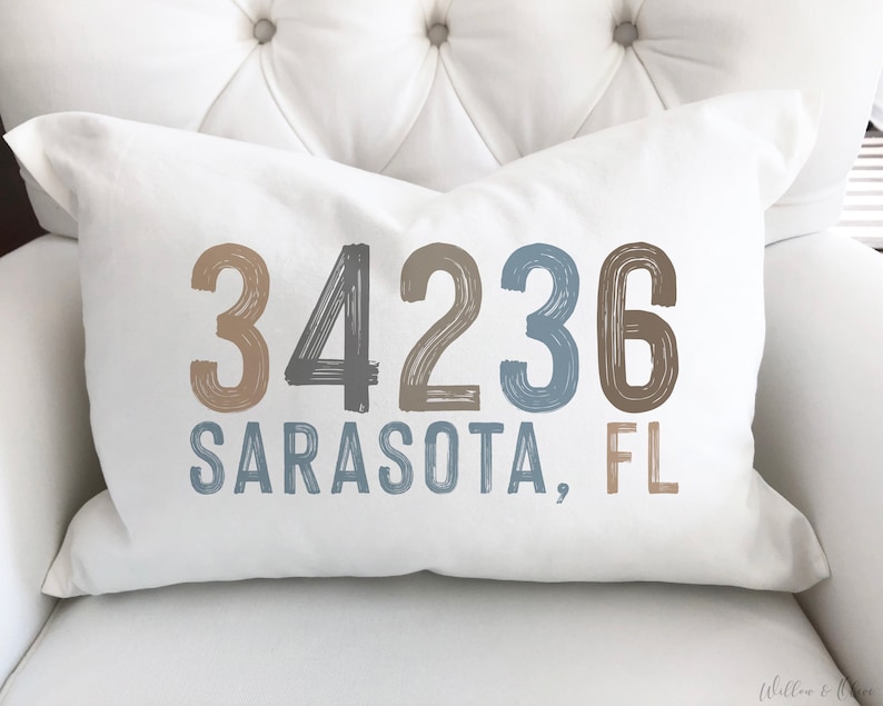 May include: White throw pillow with the text "34236 SARASOTA, FL" in a blue, gray, and brown color scheme.