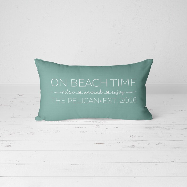 Puede incluir: Una almohada de tiro azul turquesa con texto blanco que dice "On Beach Time relax unwind enjoy The Pelican Est. 2016".