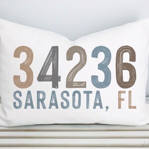 May include: White pillow with brown, gray, and blue brushstroke lettering that reads "34236 SARASOTA, FL".