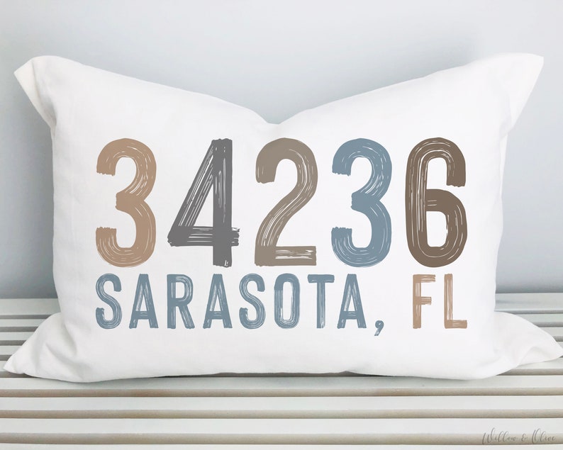 May include: White pillow with brown, gray, and blue brushstroke lettering that reads "34236 SARASOTA, FL".