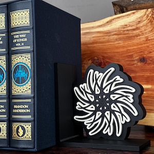 May include: Two hardcover books titled "The Way of Kings, Vol. I & II" by Brandon Sanderson, with a black bookend featuring a white and black geometric design. The books are dark blue with gold accents.